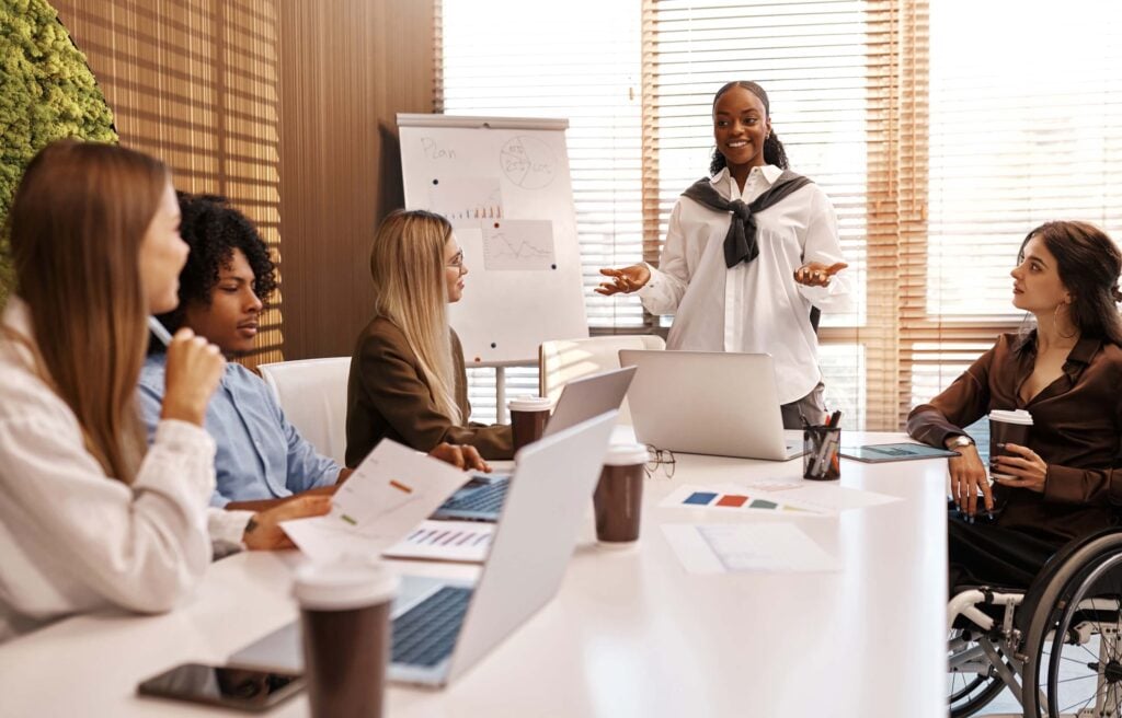 A woman stands and speaks to four colleagues, including one using a wheelchair, around a table with laptops and documents in a modern office. A flipchart with charts highlights financial management strategies in the background.