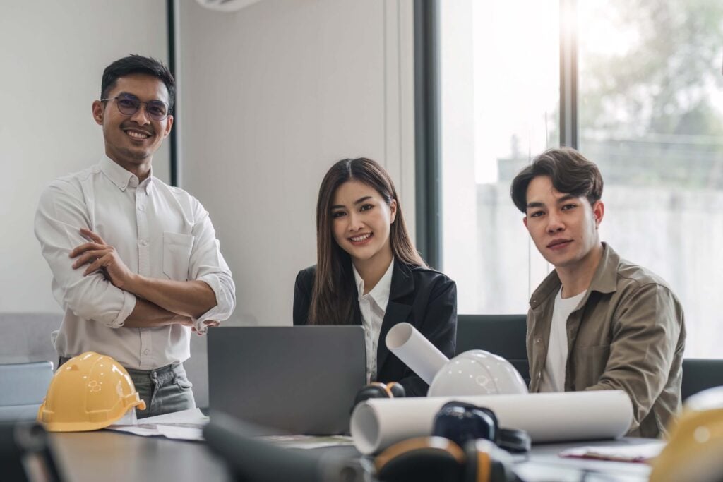 Three young professionals sit and stand around a table with blueprints, hard hats, and a laptop, smiling at the camera as they discuss a construction change directive in a bright office with large windows.