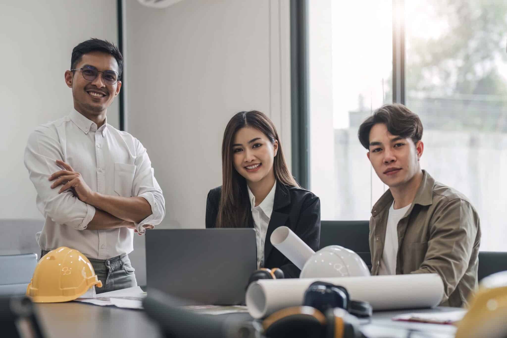 Three young professionals sit and stand around a table with blueprints, hard hats, and a laptop, smiling at the camera as they discuss a construction change directive in a bright office with large windows.