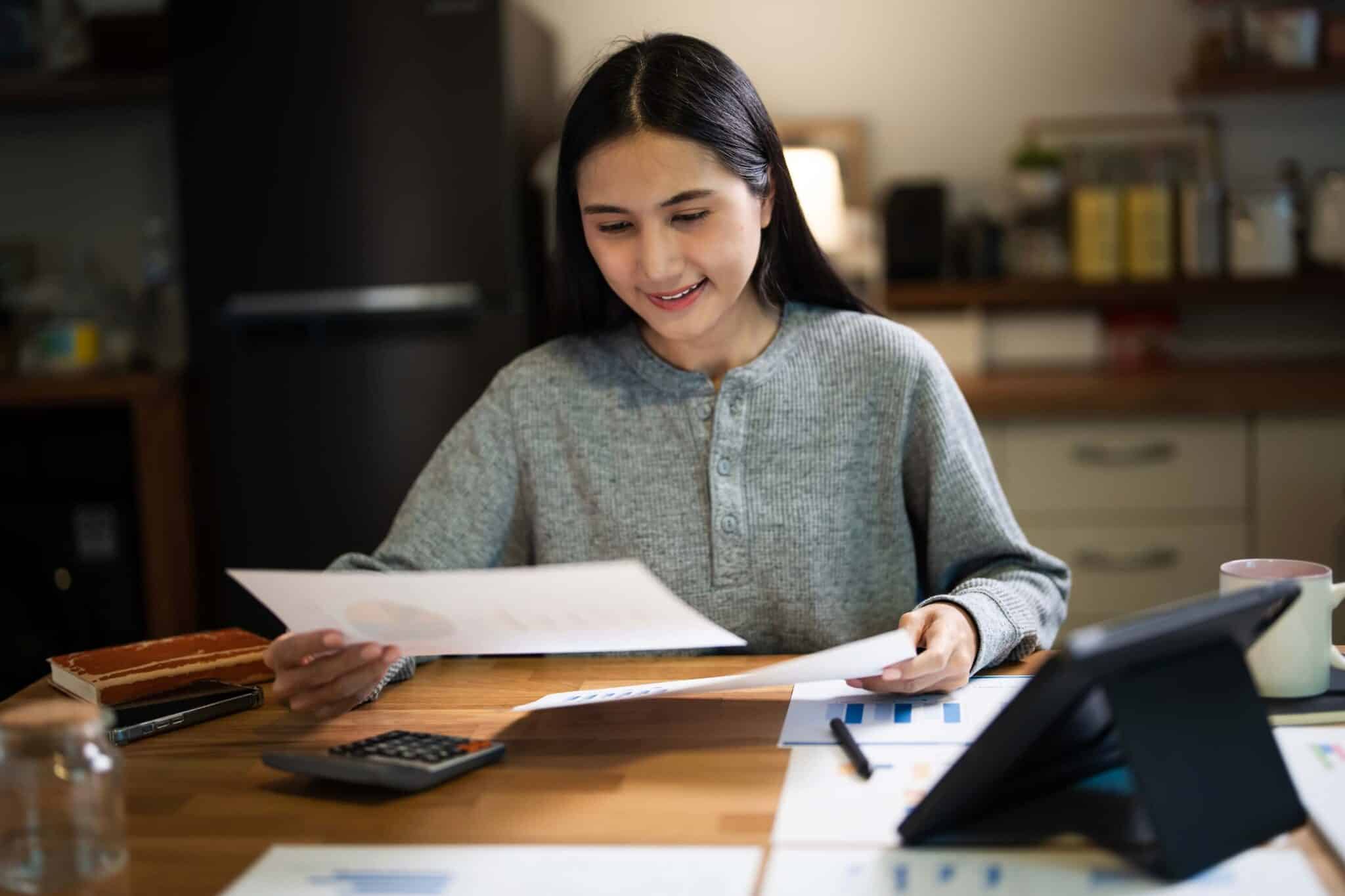 A woman sits at a table in a kitchen, smiling as she reviews title 31 documents. A tablet, calculator, mug, and various papers with charts and graphs are spread out in front of her.
