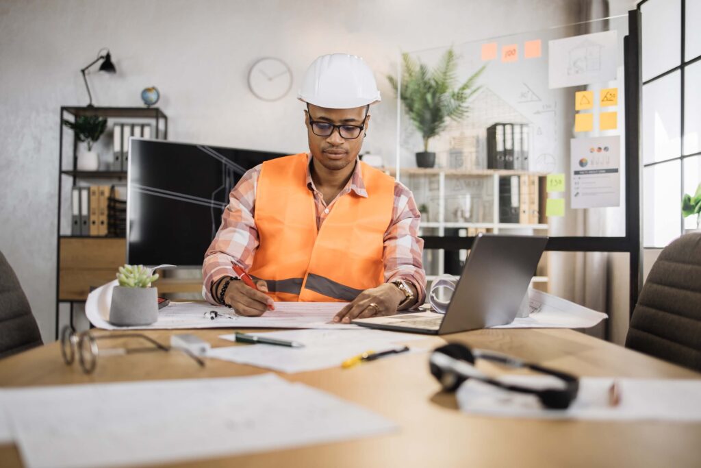 A person wearing a hard hat and orange safety vest reviews project management tasks at a desk with architectural plans, a laptop, pens, and papers in a modern office setting.
