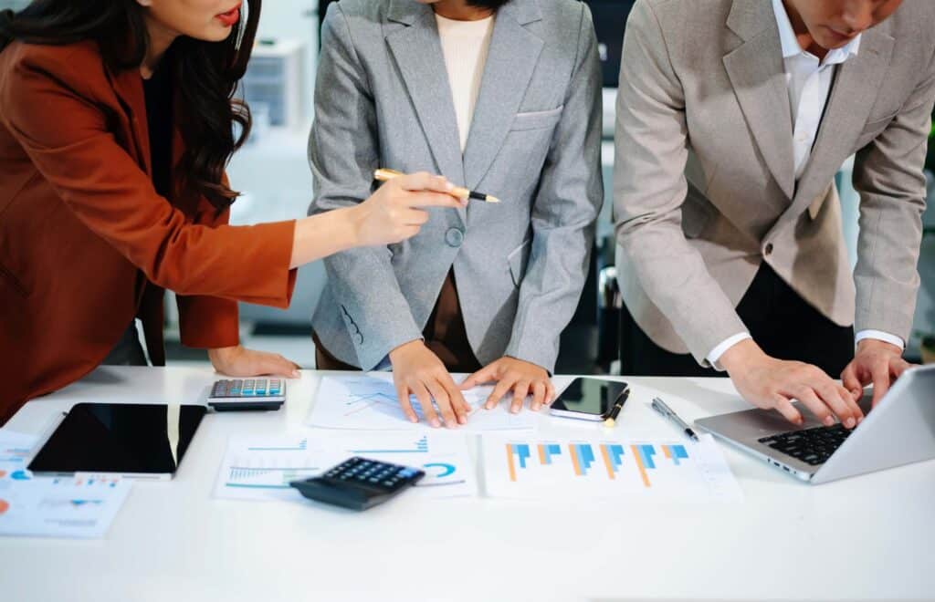 Three people in business attire work together at a desk covered with charts, graphs, a calculator, and electronic devices. One person points at a document detailing direct labor costs while another types on a laptop.