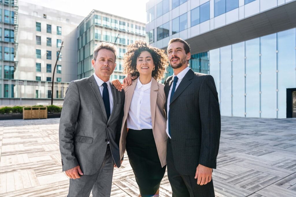 Three business professionals, two men in suits and a woman in a blazer and skirt, stand together outdoors in front of modern office buildings, smiling confidently—ready to help companies outgrowing QuickBooks reach new heights.