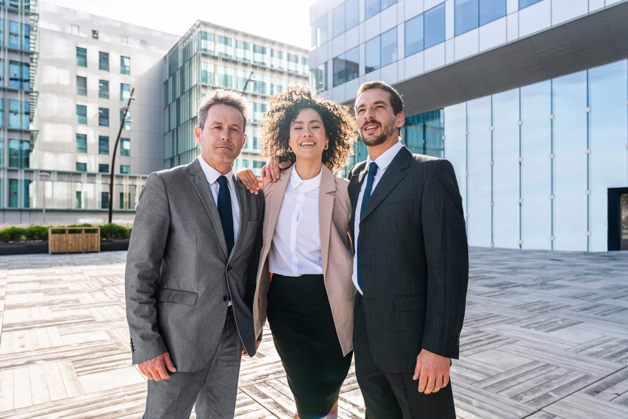 Three business professionals, two men in suits and a woman in a blazer and skirt, stand together outdoors in front of modern office buildings, smiling confidently—ready to help companies outgrowing QuickBooks reach new heights.