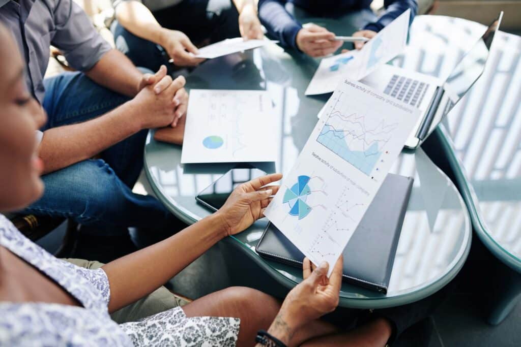 Four board members sit around a glass table with documents, charts, and a laptop, discussing printed graphs and data during a nonprofit board meeting. One person holds a sheet with diagrams for their nonprofit board report.