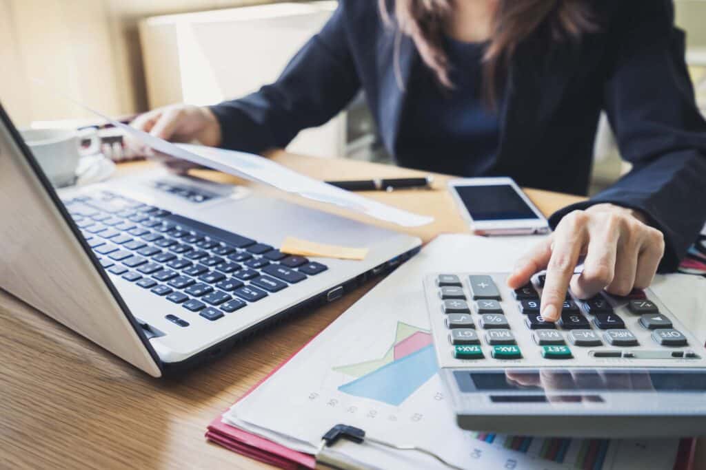A person in business attire works at a desk with a laptop, calculator, documents, and smartphone, reviewing papers and pressing calculator buttons to analyze direct labor costs, surrounded by financial charts and office supplies.
