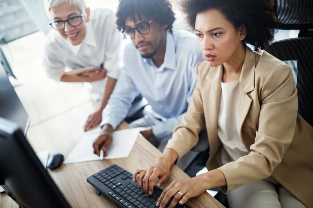 Three people collaborate at a desk; one woman types on a keyboard while two colleagues watch the computer screen and take notes. They appear focused and engaged in a bright office setting.