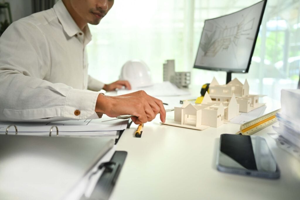 A person in a white shirt works at a desk with architectural models, papers, a smartphone, and a computer monitor displaying building plans and project progress. A white hard hat is visible in the background.