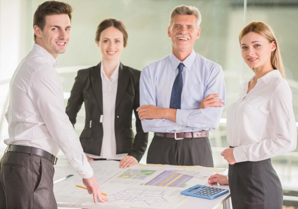 Four professionally dressed people stand around a table with blueprints and documents, smiling at the camera. One person holds a pencil, another has a calculator, as they discuss project costs and net multiplier at their business meeting.