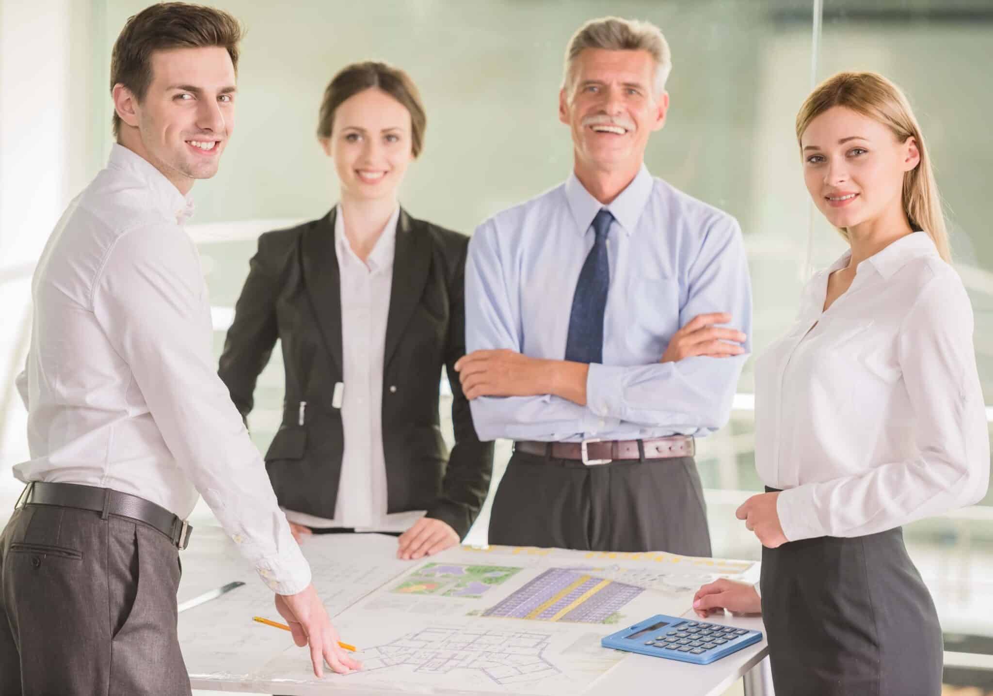 Four professionally dressed people stand around a table with blueprints and documents, smiling at the camera. One person holds a pencil, another has a calculator, as they discuss project costs and net multiplier at their business meeting.