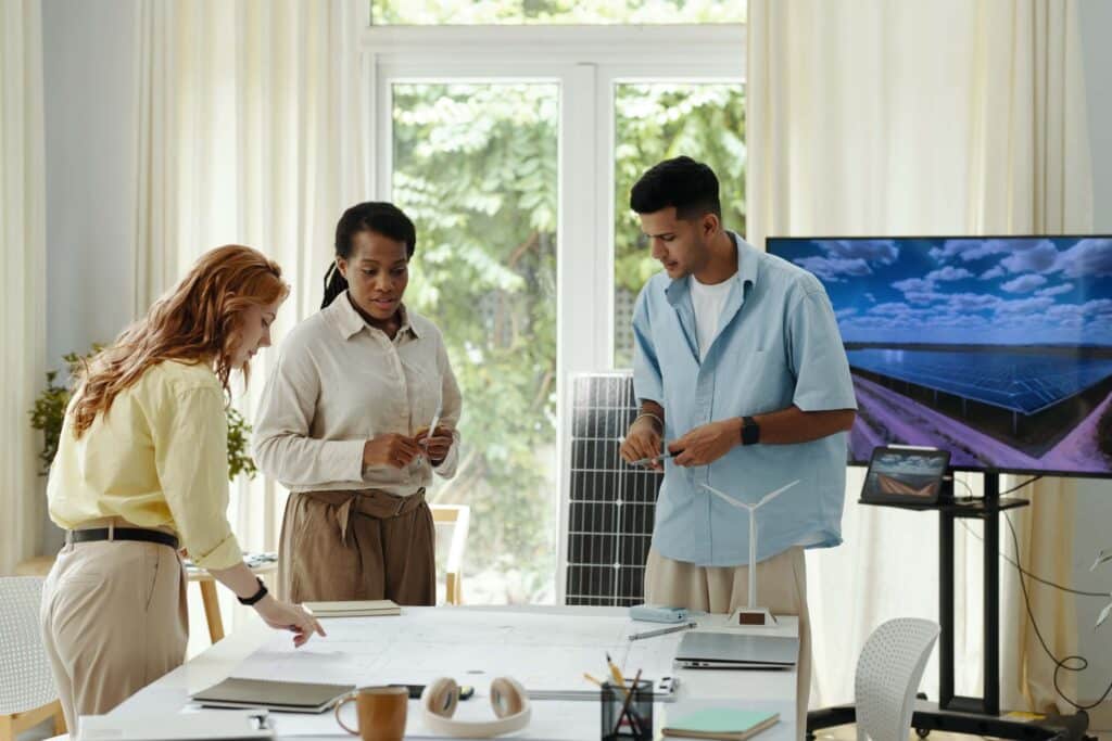 Three people collaborate around a table with blueprints, wip reports, and a model wind turbine. A solar panel and screen showing solar panels are in the background, suggesting a renewable energy project meeting.