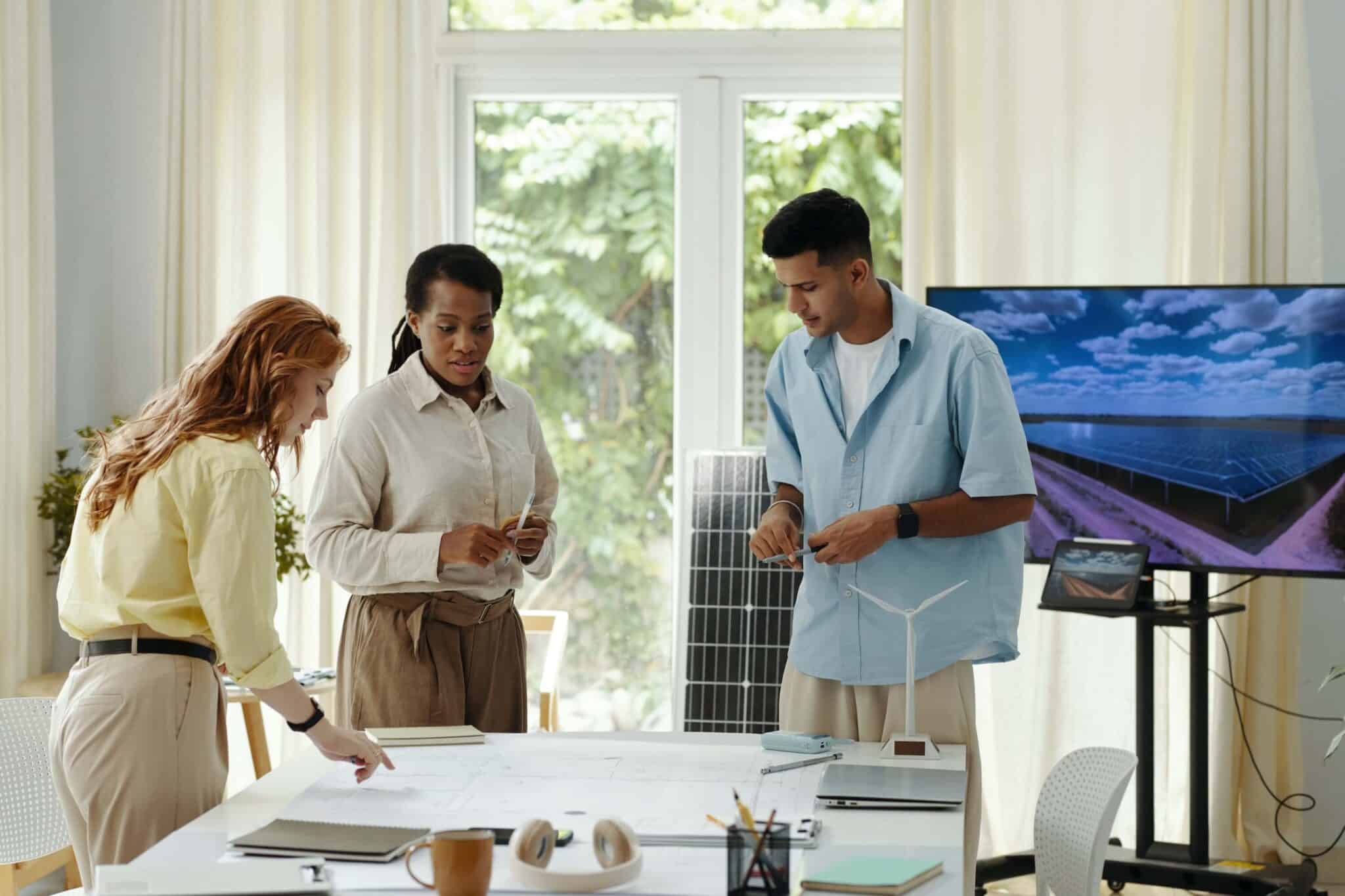 Three people collaborate around a table with blueprints, wip reports, and a model wind turbine. A solar panel and screen showing solar panels are in the background, suggesting a renewable energy project meeting.
