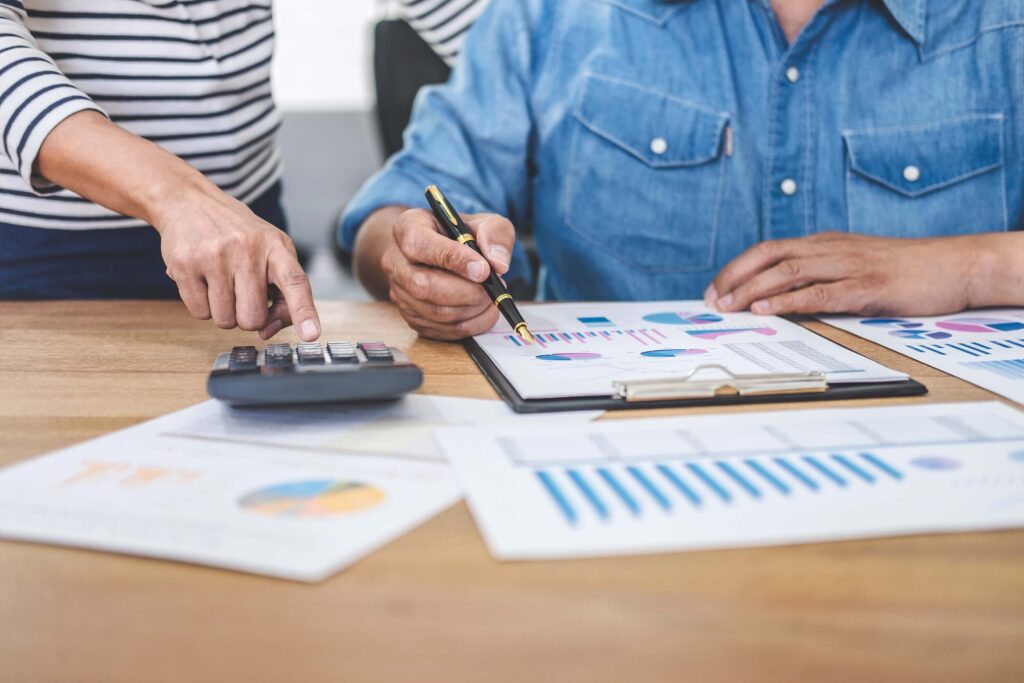 Two people analyze financial charts and graphs at a wooden desk. One points at a calculator while the other, reviewing financial statements on a clipboard, discusses the organization's financial health using various printed reports spread out on the table.