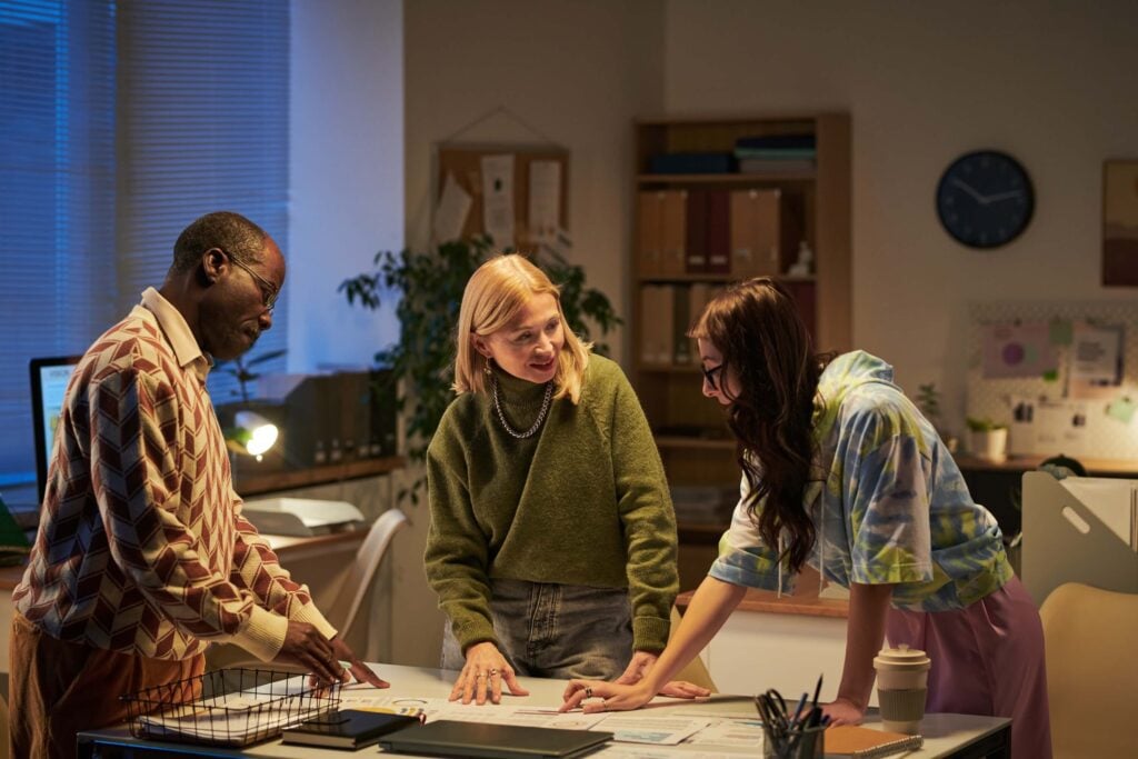 Three people stand around a desk in an office, reviewing wip reports and discussing project progress. The room’s shelves, clock, and plants create a collaborative, professional atmosphere perfect for project management meetings.