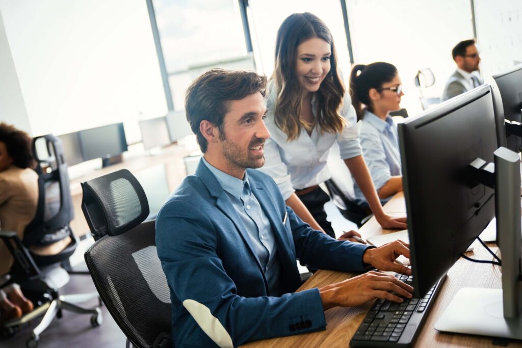 Two coworkers in business attire smile while working together at a computer, comparing Sage 100 vs Sage Intacct, in a modern, bright office space. Others are working at desks in the background.