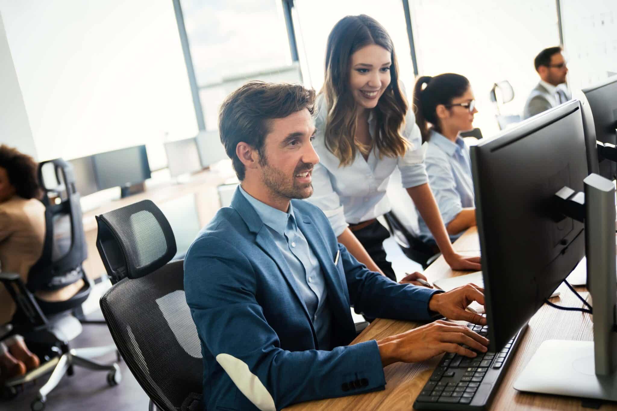 Two coworkers in business attire smile while working together at a computer, comparing Sage 100 vs Sage Intacct, in a modern, bright office space. Others are working at desks in the background.