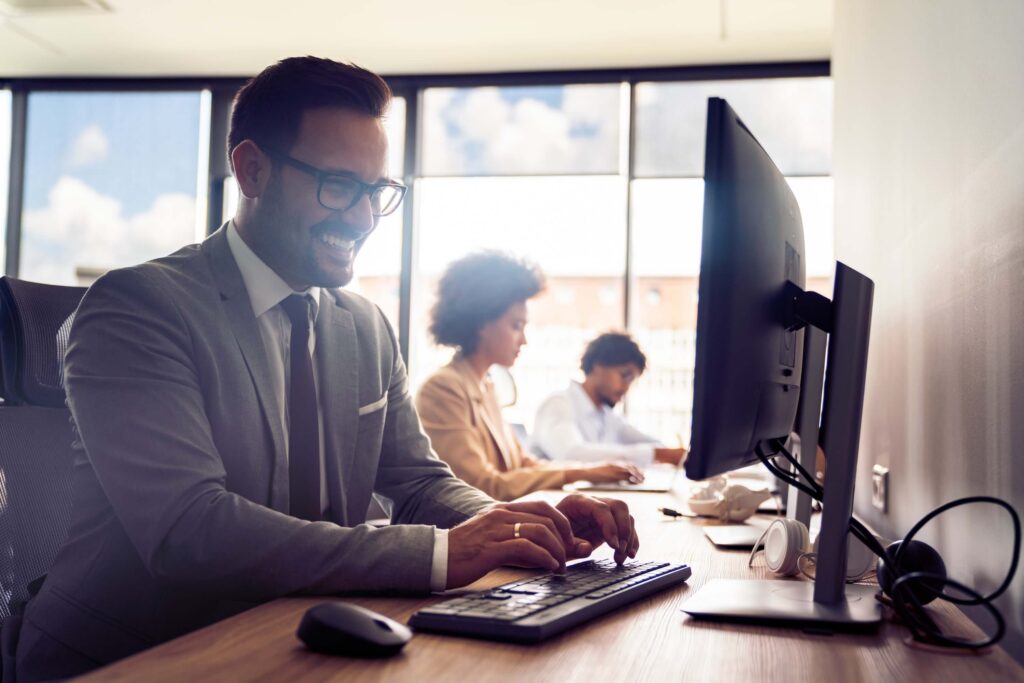 A man in a suit and glasses smiles while working on a computer at a desk, perhaps comparing Sage 100 vs Sage Intacct to optimize business processes. Two colleagues work nearby in a modern office with large windows.