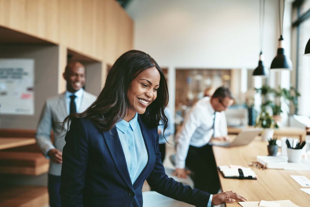 A smiling woman in a blue suit stands at a desk in a modern office, where colleagues discuss business processes. The office has large windows, natural light, and a professional atmosphere—ideal for conversations about Sage 100 vs Sage Intacct solutions.