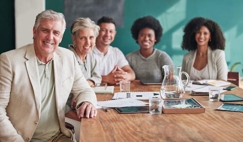 Five people of diverse ages and backgrounds sit together at a conference table, smiling at the camera during a nonprofit board report. Papers, tablets, a water pitcher, and glasses are on the wooden table in front of them.