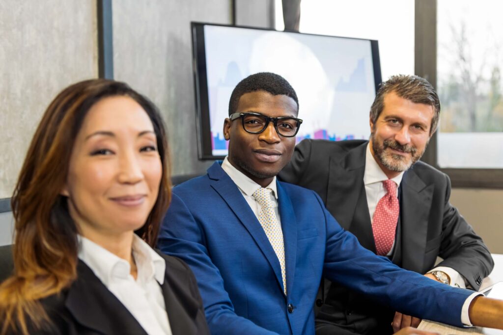 Three business professionals, two men and one woman, sit together at a conference table. They are dressed in formal business attire, smiling, with a large screen and windows in the background.