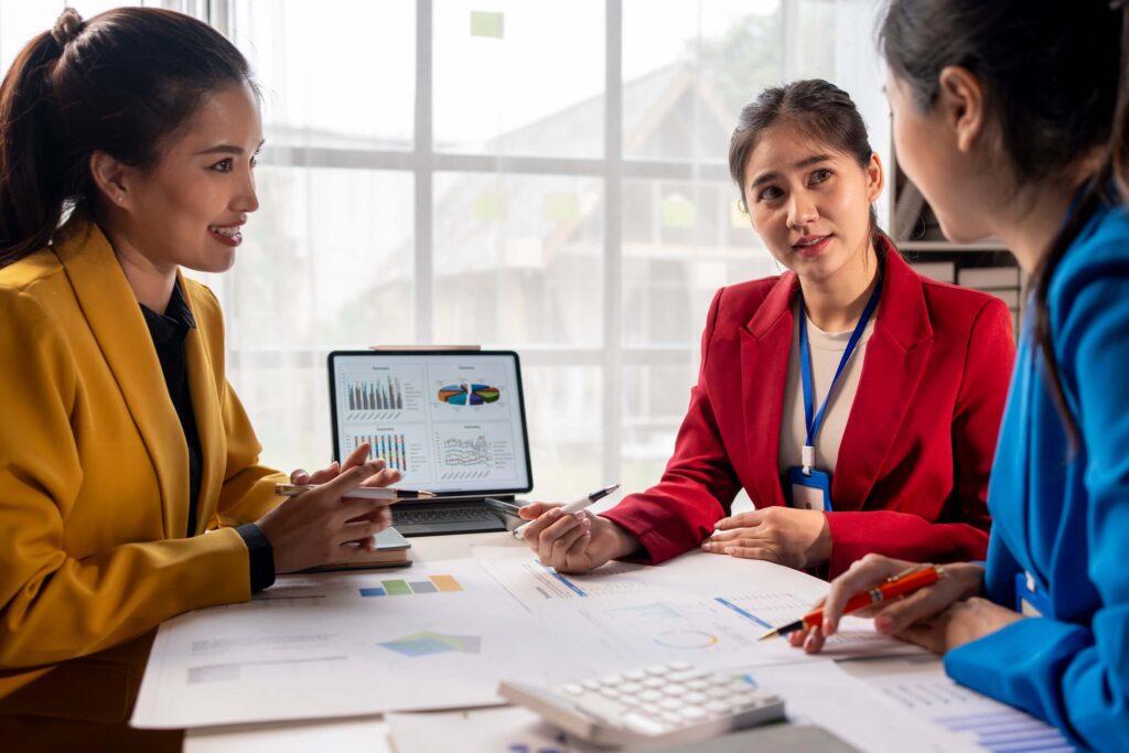 Three women in business attire sit at a table discussing financial statements and charts, with a laptop displaying graphs in the background. They appear engaged in a collaborative meeting focused on their organization's financial health.