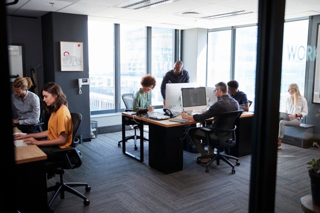 Modern office with large windows, six people working at desks and computers—some collaborating on business processes, others focusing individually; bright, spacious, and professional atmosphere.