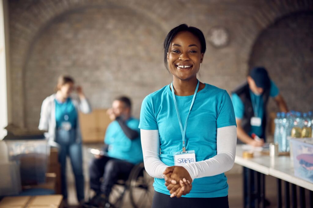 A smiling woman wearing a blue volunteer shirt and name tag stands in the foreground, with people packing supplies behind her at a community service event supporting the organization's financial health.