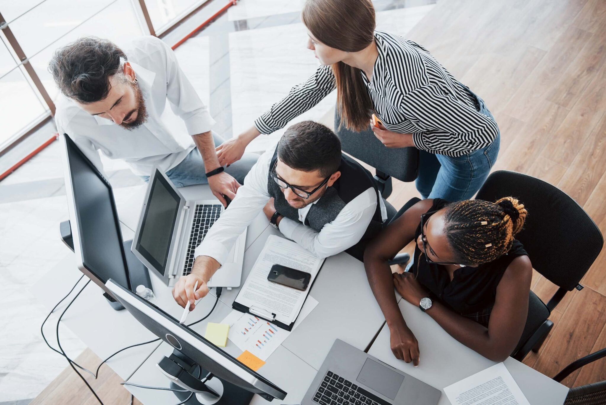 Four people collaborate at a desk with computers, laptops, and papers. One points at a monitor while others discuss a Sage Intacct implementation, highlighting teamwork in an office environment.