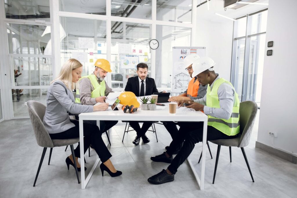 Five people, including two in suits and three in construction gear with hard hats and vests, sit at a table in a modern office, reviewing documents and plans during a meeting focused on project management.