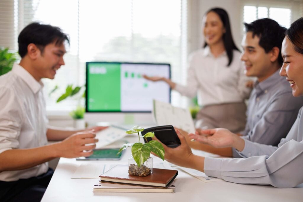 Four people in business attire sit around a table with documents, a plant, and a tablet, while a woman stands and gestures toward a presentation on a screen in a bright office. The group discusses important topics such as Sage 100 End of Life and how to handle reports from the closed fiscal year, following just a general guideline for best practices.