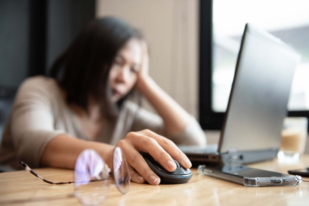 A person sits at a desk, using a computer mouse with a laptop in front of them. Glasses and a pen are on the table, and the person appears tired or bored with their hand holding their head—perhaps feeling overwhelmed after reviewing reports from a recently closed fiscal year.