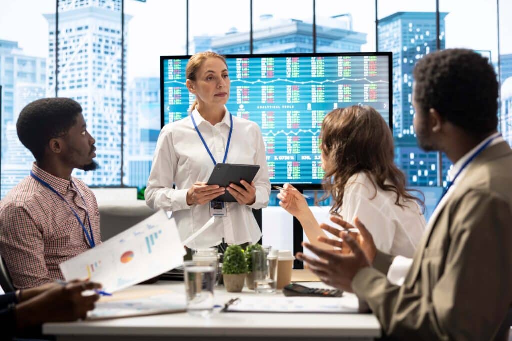 A woman holding a tablet speaks to four colleagues in a modern office. Stock market data and fund accounting charts are displayed on a large screen behind her as the group listens and discusses graphs and analytics.