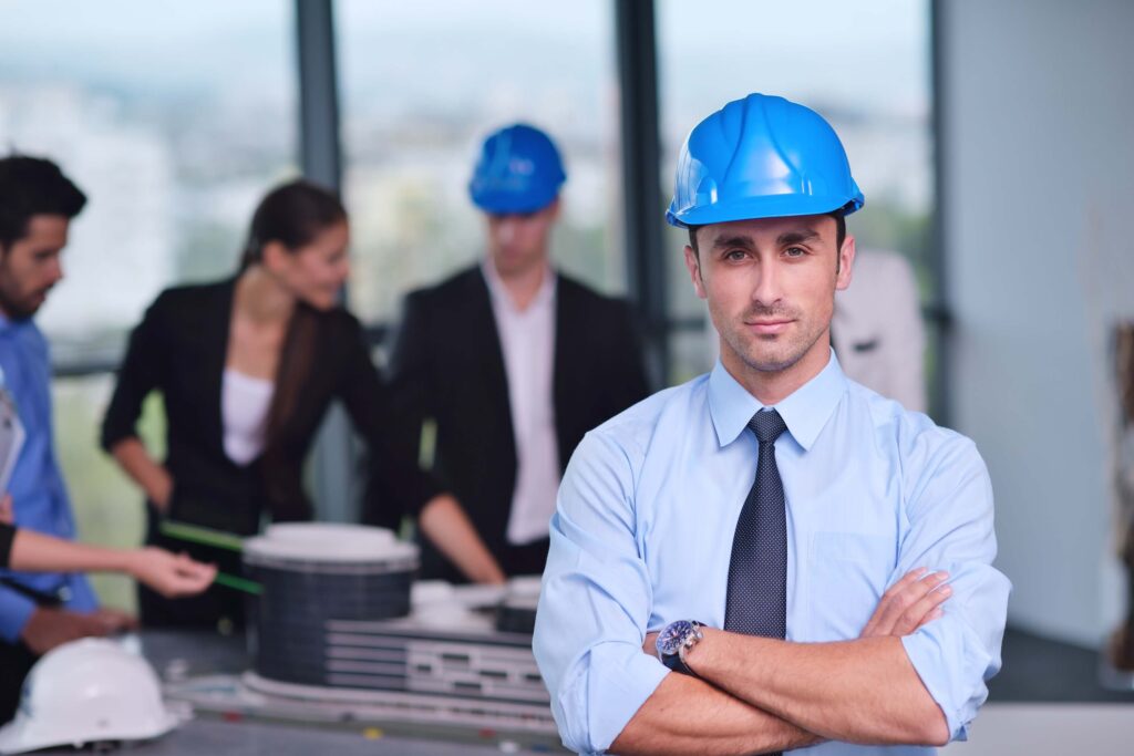 A man in a blue hard hat and dress shirt stands with arms crossed, while three people in business attire and hard hats discuss a project at a table in the background, illustrating an effective project management environment enhanced by Procore's project management platform.