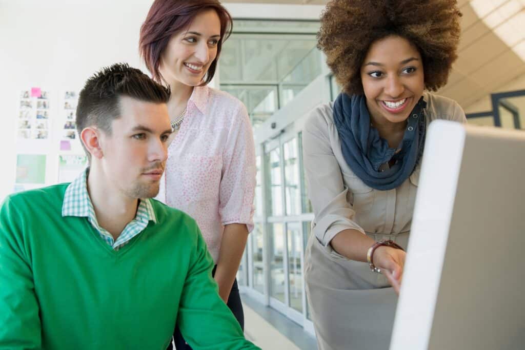 Three people gather around a computer in a bright office. One woman points at the monitor, while the other two, a man and woman, watch and smile, appearing engaged in a collaborative discussion about financial data and utilizing Sage Intacct dimensions to streamline their workflow.