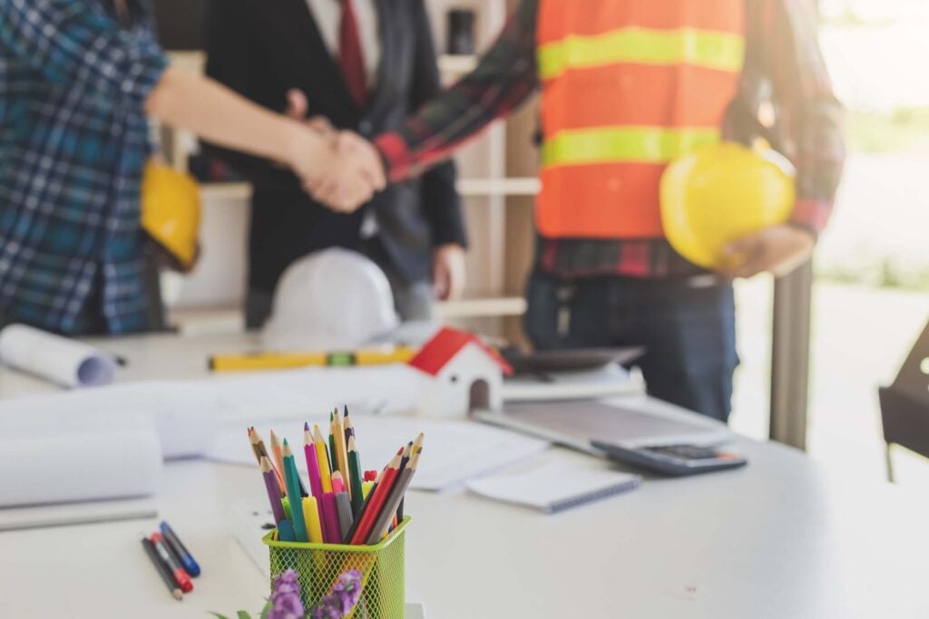 Two people are shaking hands over a desk with construction plans and supplies, highlighting a successful collaboration powered by Procore's project management platform. One person wears a plaid shirt and the other wears a safety vest and holds a yellow helmet, emphasizing the teamwork essential on site. Pencils and papers are in the foreground, representing organized planning enhanced by seamless Procore integrations.