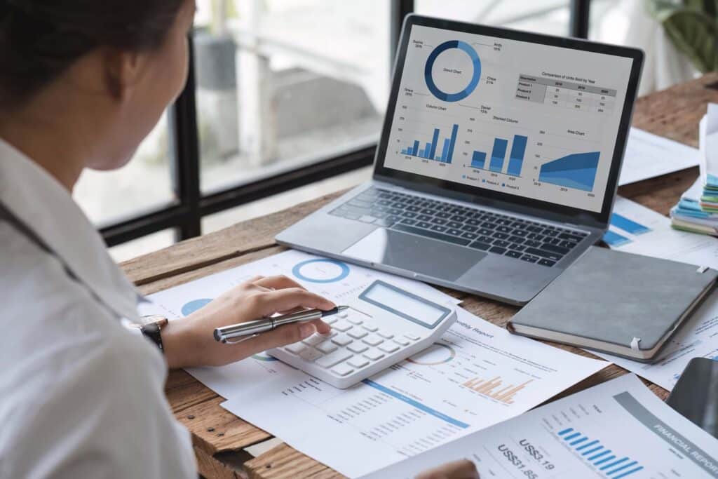 A person uses a calculator and reviews financial graphs and charts on paper and a laptop at a wooden desk with reports, a notebook, and a pen, analyzing financial data using Sage Intacct dimensions to gain deeper insights.