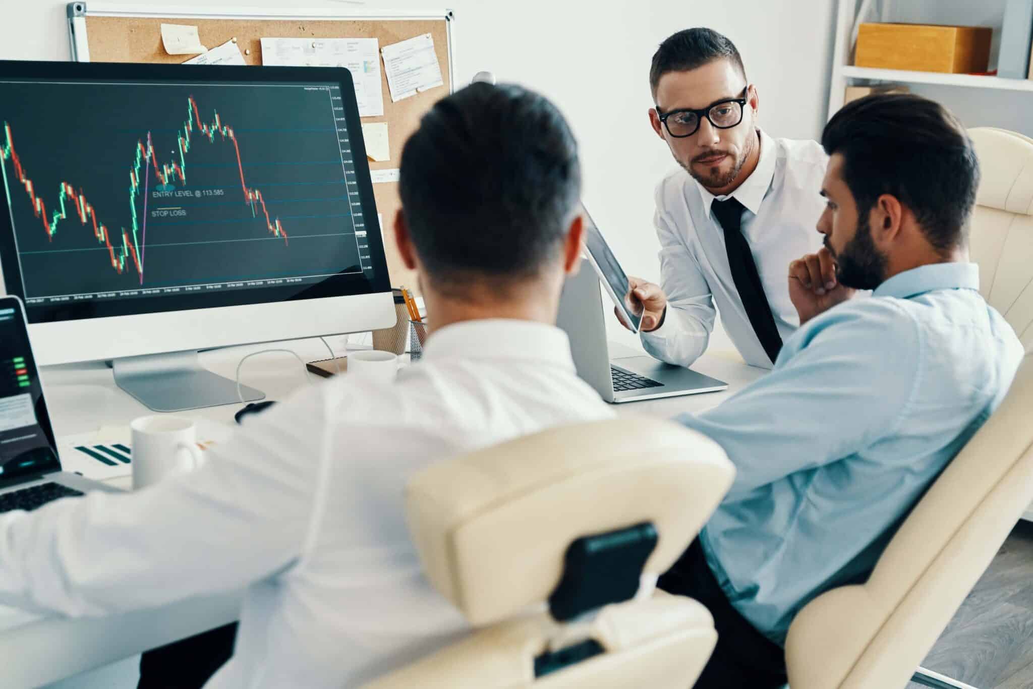 Three men in business attire sit at a desk in an office, discussing financial data on a computer monitor showing a declining stock chart, while private equity accounting software is open on their laptops amidst papers and office supplies.