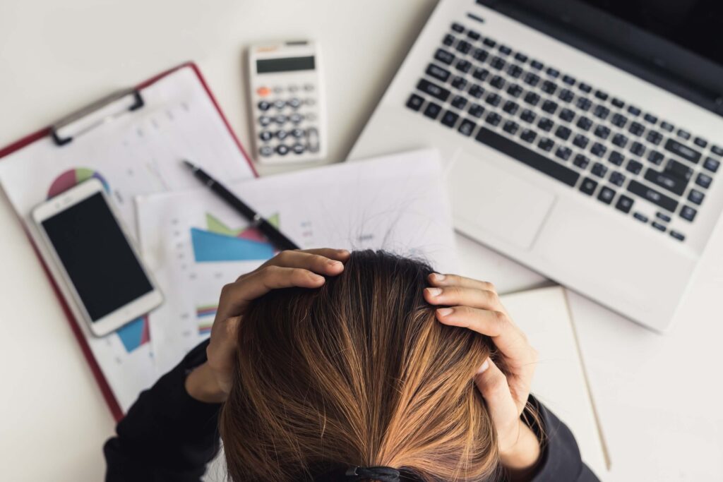 A person holds their head in frustration at a desk with a laptop, smartphone, calculator, and financial documents, suggesting stress over work or finances—possibly overwhelmed by complex financial data or navigating dimensional accounting in Sage Intacct.