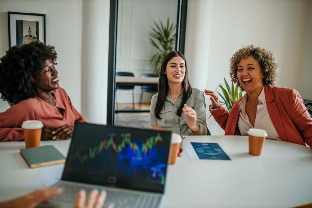 Three women sit around a table with coffee cups, smiling and talking. A blurred laptop screen displays a colorful fund accounting chart in the foreground. The atmosphere appears casual and friendly.