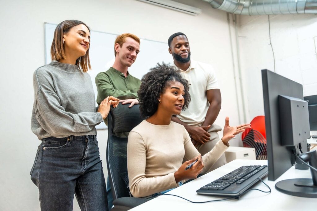 Four people are gathered around a desk, looking at a computer screen. One woman is seated and gesturing while the others stand nearby, appearing engaged and interested in the discussion about optimizing business processes through Deltek Vantagepoint integrations.