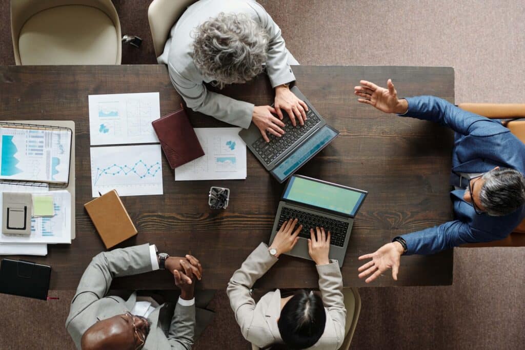 Four people sit around a table with laptops, documents, and charts, engaged in a business meeting about private equity accounting software. One person gestures while others work on laptops and review papers. The scene is viewed from above.