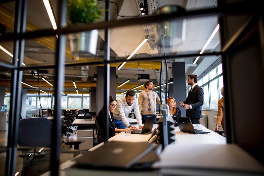 Six people in a modern office having a discussion—some seated at desks with laptops and others standing—are viewed through a black metal shelf. This bright, open workspace with large windows fosters collaboration as the team explores ways to streamline business processes and improve data synchronization across their projects.