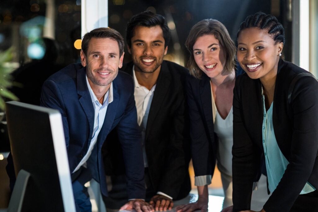 Four professionally dressed people, two men and two women, smiling as they gather around a desk in an office at night, with city lights visible through the window behind them. Whether they're discussing a closed fiscal year or following just a general guideline for project management, their collaborative spirit is evident in this professional setting.