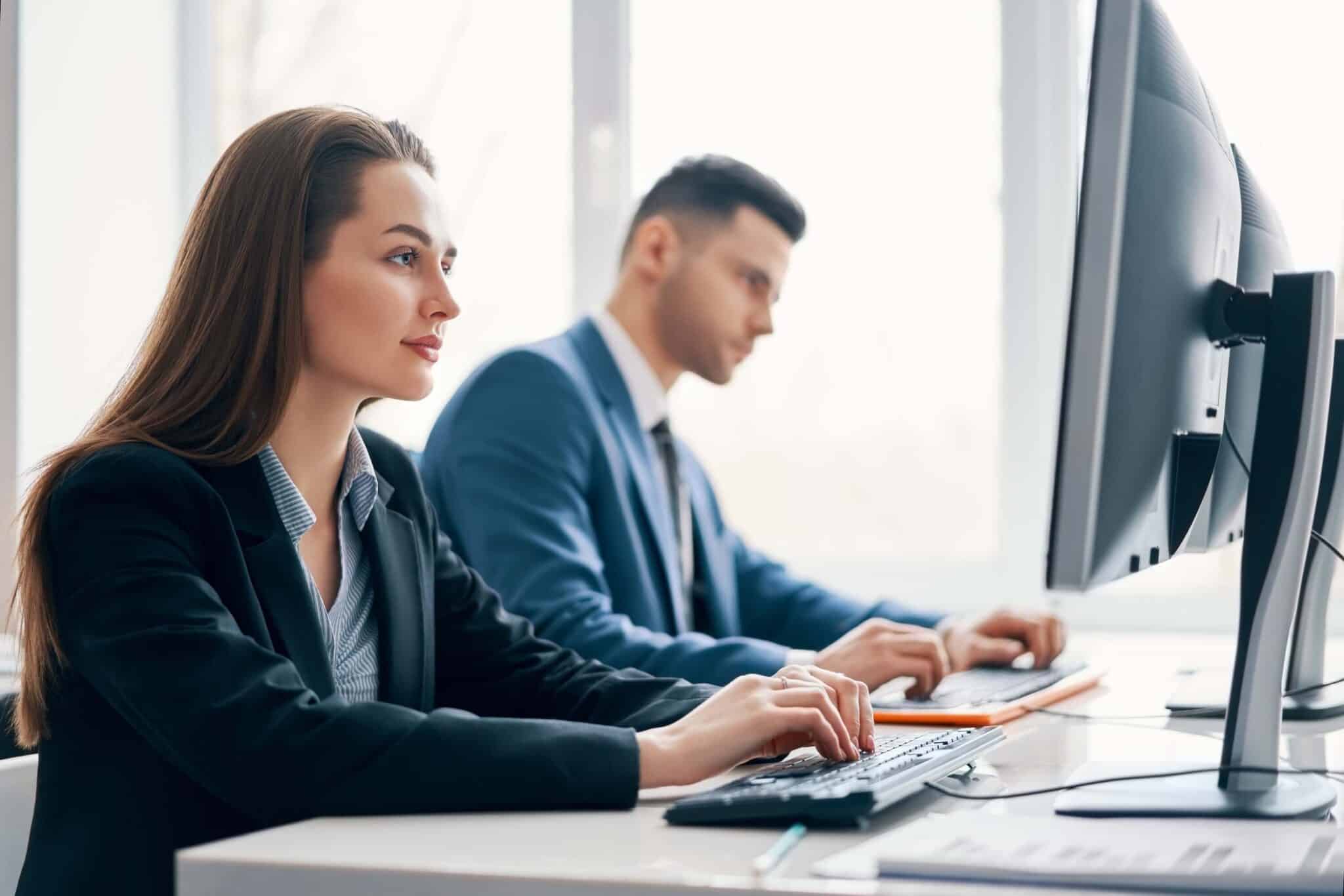 Two professionals in business attire work at desktop computers in a bright, modern office. The woman in the foreground types while focusing on dimensional accounting in Sage Intacct, while the man in the background concentrates on his screen.
