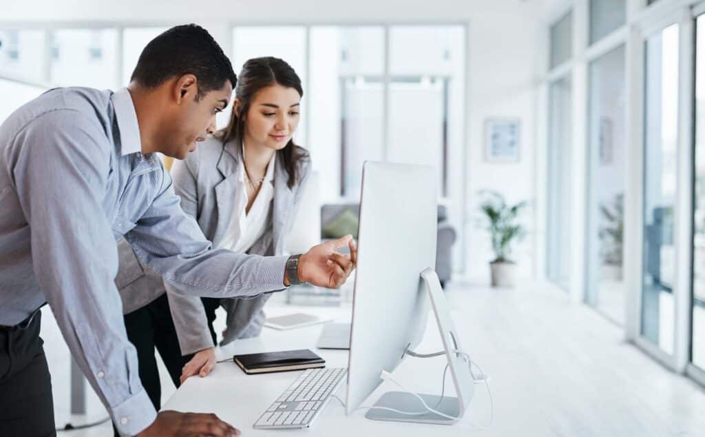 Two professionals in business attire look at a computer monitor together in a bright office. One points at the screen while the other observes attentively, as they review financial data using Sage Intacct dimensions. A notebook and phone are on the desk.