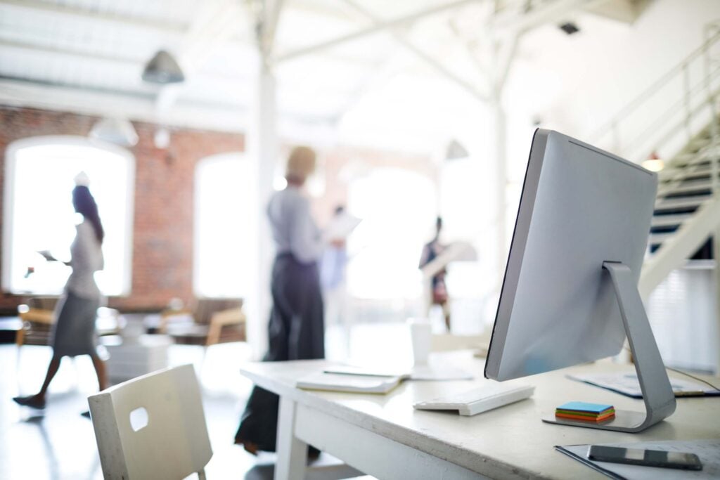 A modern, bright office with a computer monitor on a desk in the foreground—displaying seamless data synchronization—and blurred people working and talking in the background, all collaborating to enhance business processes.