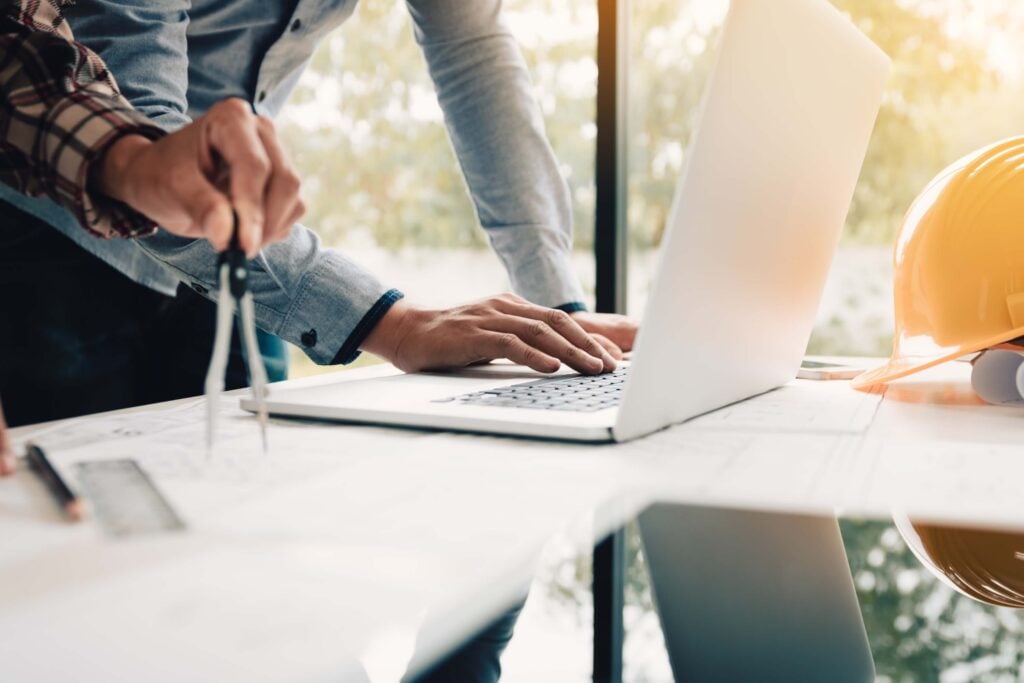 Two people work at a desk with building plans, one using a laptop—perhaps reviewing details on the Deltek Vantagepoint mobile app—and the other holding a compass. A yellow construction helmet sits nearby, and sunlight streams through a window.