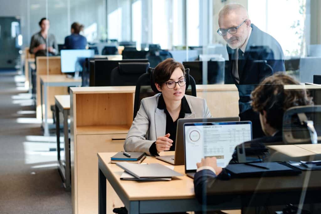 Three professionals confer at a modern office desk. One person shows data on a laptop, while two others listen attentively—reviewing financial insights powered by Beanworks by Quadient. Open workspaces, papers, and glass dividers are visible in the background.