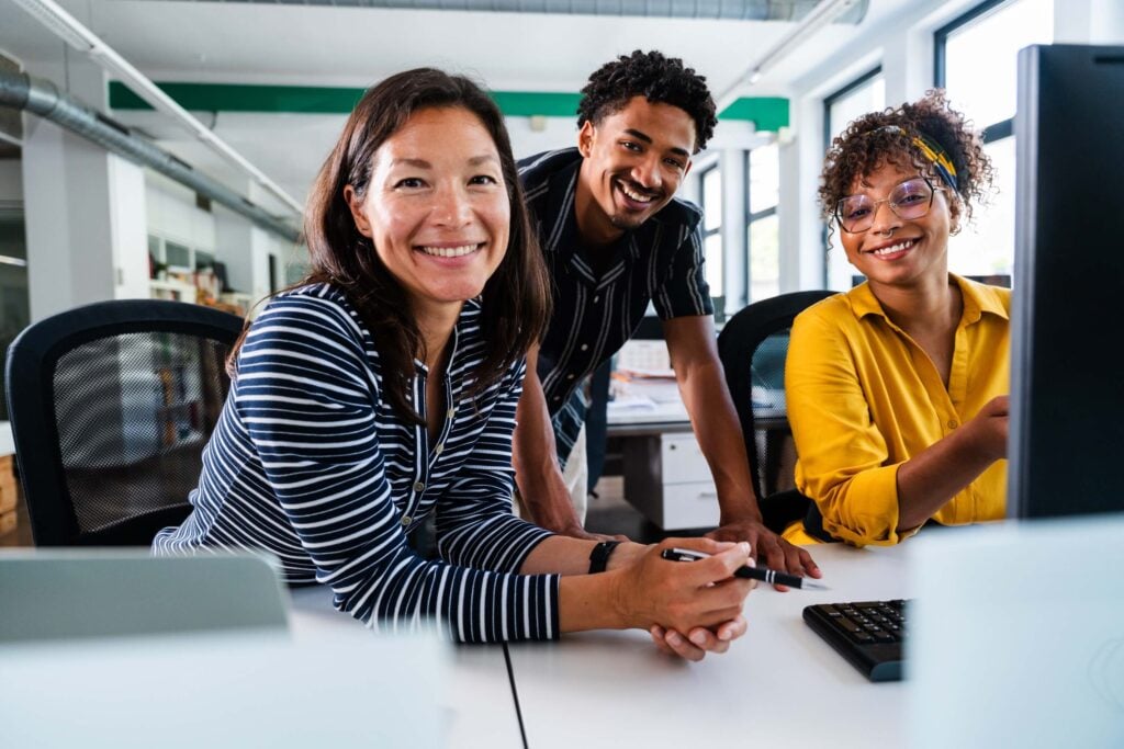 Three colleagues sit and stand around a desk in a bright office, smiling at the camera, with a computer and paperwork visible on the desk—perhaps reviewing new solutions from the Sage Intacct Marketplace to enhance their workflow.