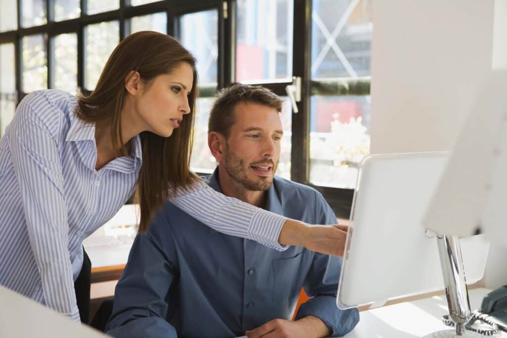 A woman stands beside a seated man, both looking at a computer screen displaying Deltek Vantagepoint reports. The woman points at the monitor, and they appear to be discussing something. Sunlight streams in from large windows behind them.
